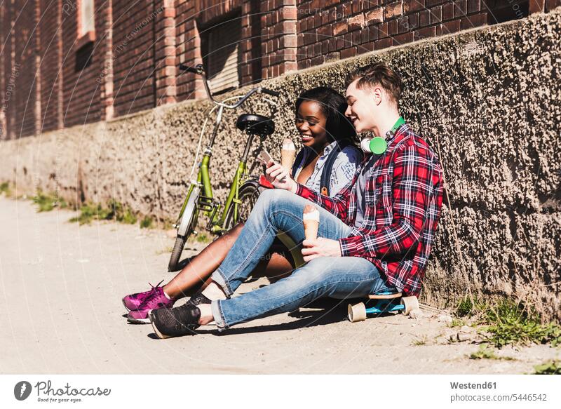 Junges Paar sitzt am Boden und isst Eiscreme jung multikulturell flirten Flirt Pärchen Paare Partnerschaft Handy Mobiltelefon Handies Handys Mobiltelefone essen