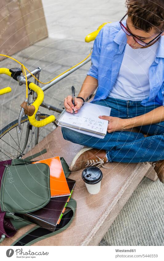 Junger Mann mit Rennrad sitzt auf Bank und schreibt auf Notizblock Student Hochschueler Studierender Hochschüler Studenten schreiben aufschreiben notieren
