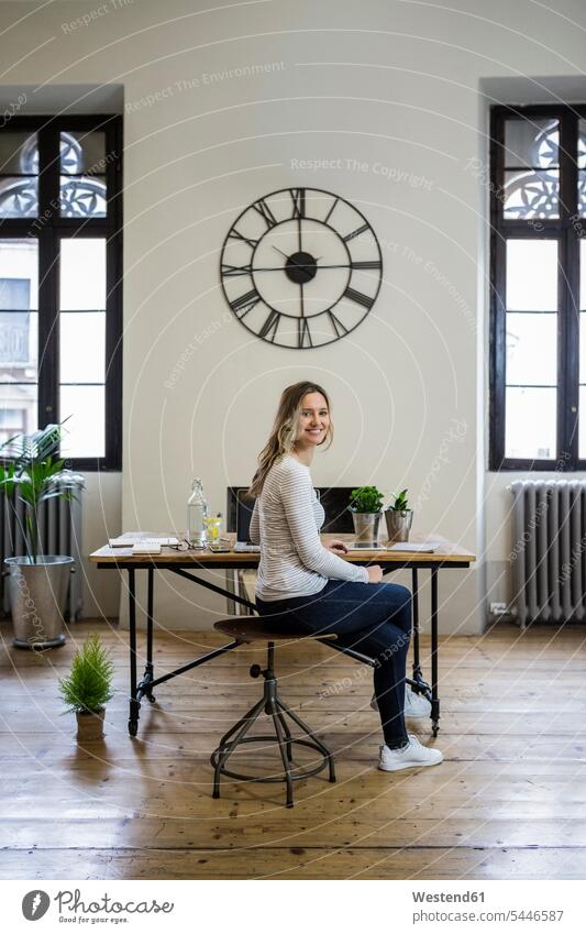 Porträt einer lächelnden Frau, die zu Hause am Schreibtisch unter einer großen Wanduhr sitzt Arbeitstisch Schreibtische Wanduhren Portrait Porträts Portraits