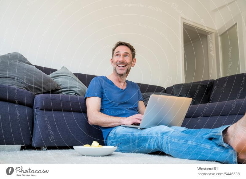 Porträt eines lachenden Mannes mit Laptop, der im Wohnzimmer auf dem Boden sitzt Männer männlich Notebook Laptops Notebooks Erwachsener erwachsen Mensch
