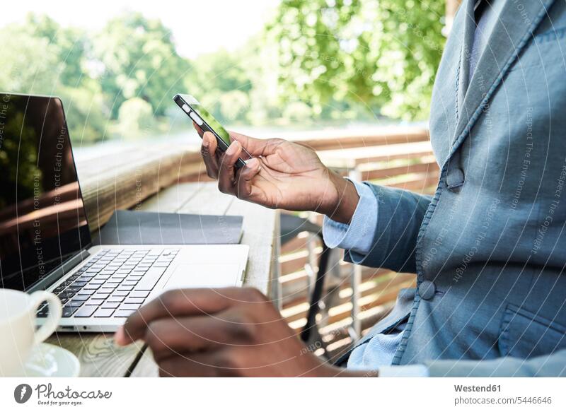 Geschäftsmann überprüft Telefon, während er mit seinem Laptop auf einer Terrasse arbeitet, Teilansicht Smartphone iPhone Smartphones Hand Hände Businessmann