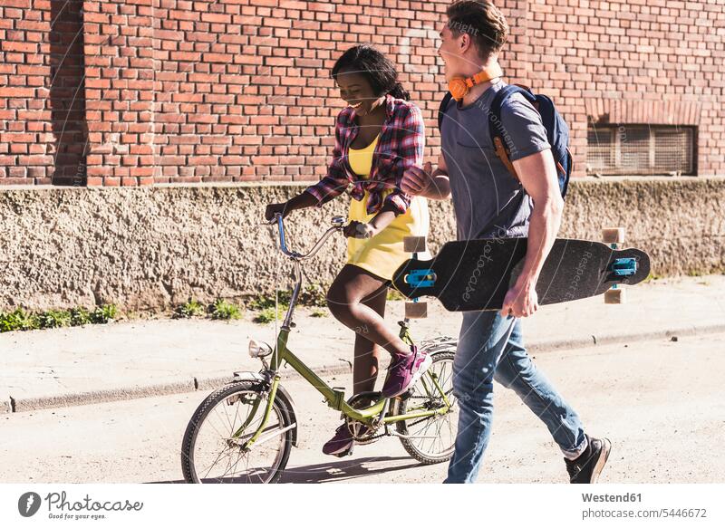 Junges Paar mit Fahrrad und Skateboard auf der Straße multikulturell Bikes Fahrräder Räder Rad gehen gehend geht Strassen Straßen jung flirten Flirt Rollbretter
