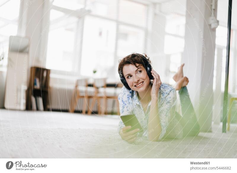 Frau, die in ihrer Wohnung auf dem Boden liegt und ein Smartphone und Kopfhörer benutzt hören hoeren weiblich Frauen Kopfhoerer Musik Fußboden Fußboeden