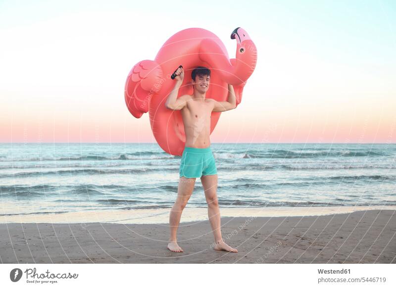 Lächelnder junger Mann mit aufblasbarem rosa Flamingo am Strand bei Sonnenuntergang Beach Straende Strände Beaches Männer männlich Erwachsener erwachsen Mensch