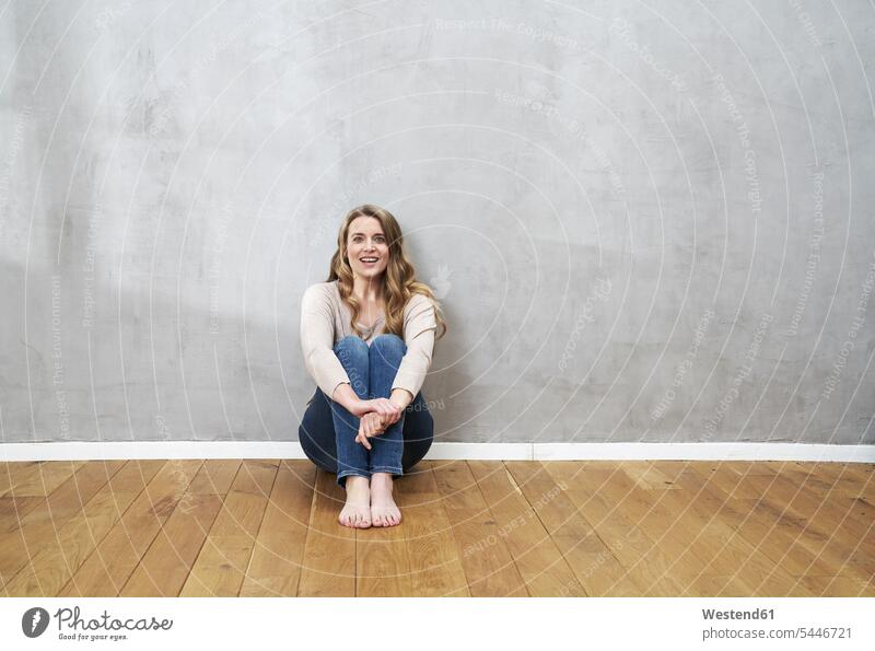Lächelnde blonde Frau sitzt auf dem Boden vor grauer Wand Portrait Porträts Portraits weiblich Frauen Erwachsener erwachsen Mensch Menschen Leute People