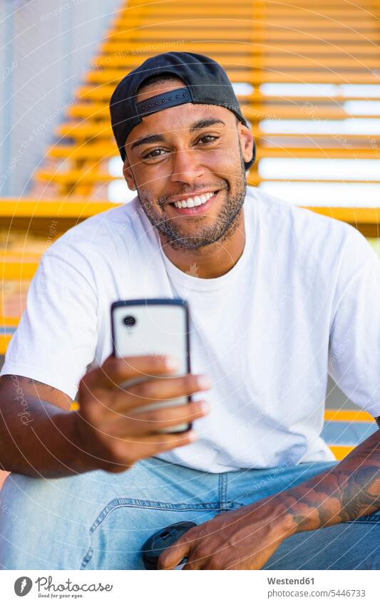 Porträt eines lächelnden jungen Mannes mit Smartphone, der auf einer Treppe sitzt Portrait Porträts Portraits iPhone Smartphones Männer männlich Handy