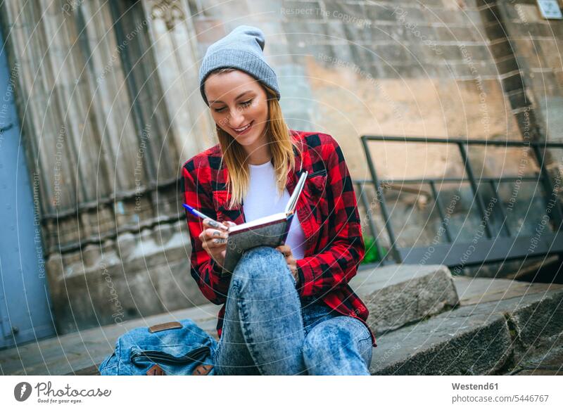 Spanien, Barcelona, lächelnde junge Frau sitzt auf einer Treppe und schreibt in ein Notizbuch weiblich Frauen Notizbücher Notizbuecher Erwachsener erwachsen