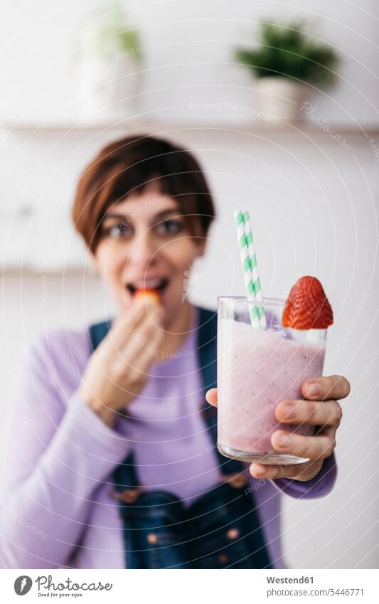 Frau hält ein Glas Erdbeer-Smoothie, während sie eine Frucht isst Smoothies Getränk Getraenk Getränke Getraenke Food and Drink Lebensmittel Essen und Trinken