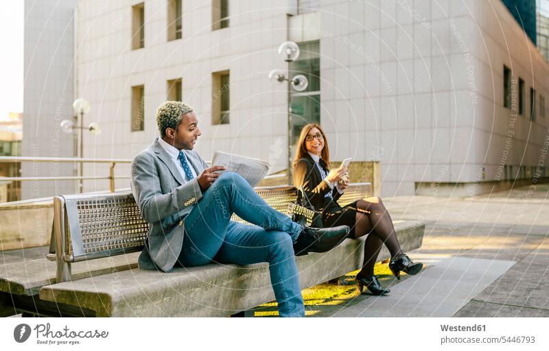 Junger Geschäftsmann und junge Geschäftsfrau sitzen auf einer Bank und unterhalten sich Geschäftsleute Geschäftspersonen unterwegs auf Achse in Bewegung