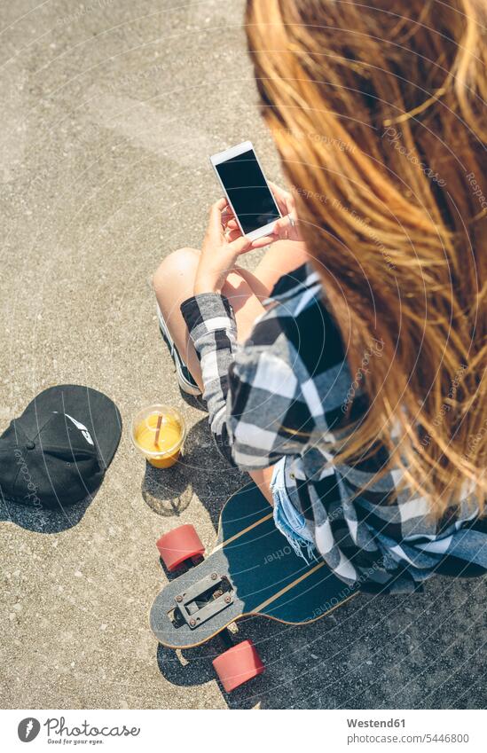 Rückenansicht einer jungen Frau mit langen Haaren, die auf einem Longboard sitzt und auf ein Smartphone schaut weiblich Frauen Erwachsener erwachsen Mensch
