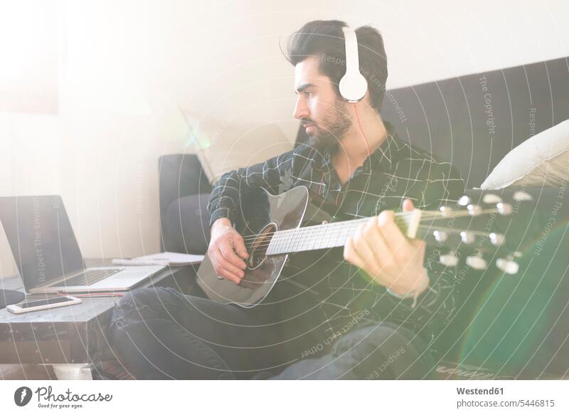 Junger Mann spielt zu Hause Gitarre und trägt Kopfhörer, die an einen Laptop angeschlossen sind Männer männlich Gitarren Notebook Laptops Notebooks Erwachsener