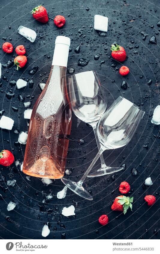 Eine Flasche Rosenwein, zwei leere Weingläser, Eiswürfel, Erdbeeren und Himbeeren auf dunklem Grund eisgekühlt eisgekuehlt eisgekühlte schwarzer Hintergrund
