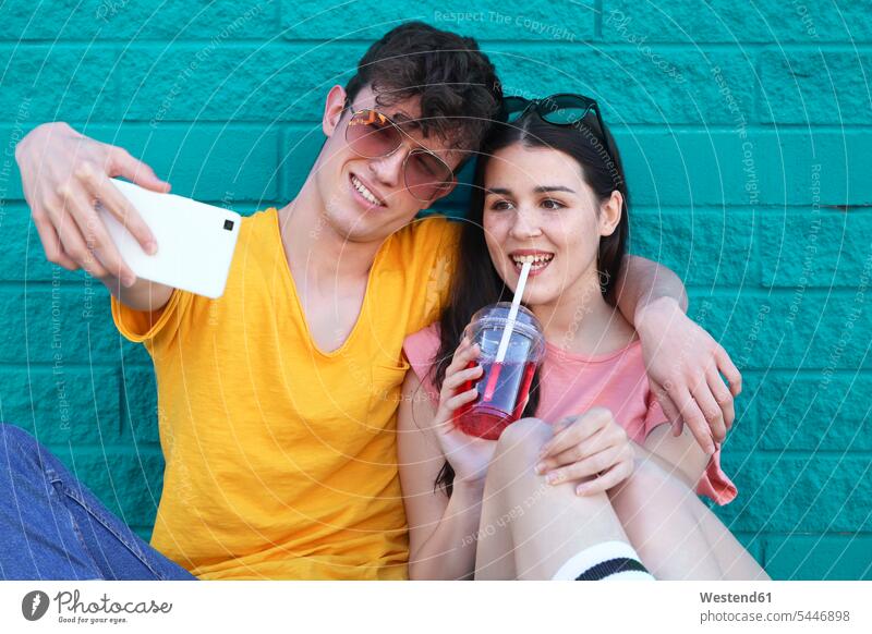 Junges Paar, das ein Selfie mit Smartphone vor einer blauen Backsteinmauer macht Selfies Pärchen Paare Partnerschaft Portrait Porträts Portraits Mensch Menschen