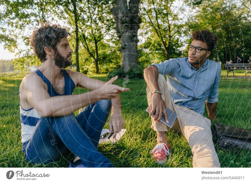 Zwei Freunde sitzen in einem Park mit mobilem Gerät und Papieren und diskutieren Mann Männer männlich sitzend sitzt Parkanlagen Parks besprechen Besprechung