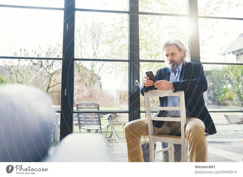 Leitender Geschäftsmann sitzt auf einem Stuhl und benutzt ein Smartphone Businessmann Businessmänner Geschäftsmänner SMS Textnachricht benutzen benützen Handy