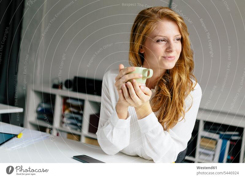 Lächelnde junge Frau mit Tasse Kaffee am Schreibtisch im Büro lächeln Zuversicht Zuversichtlich Selbstvertrauen selbstbewusst Vertrauen weiblich Frauen Office