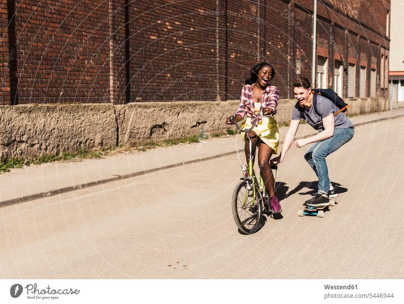 Junge Frau auf Fahrrad ziehender junger Mann, stehend auf Skateboard multikulturell Skateboarden Skateboardfahren Skateboarding Rollbretter Skateboards Paar