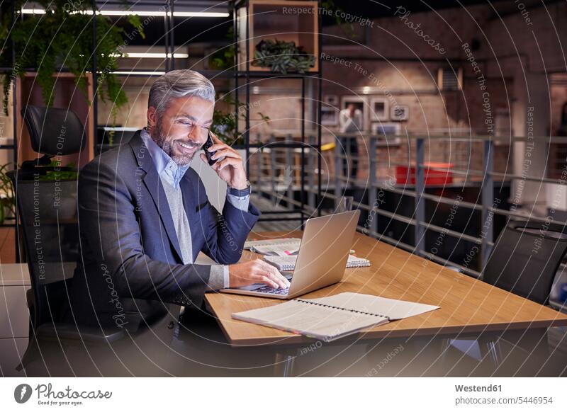 Ein reifer Geschäftsmann, der in einem modernen Büro arbeitet und während des Telefonierens einen Laptop benutzt sitzen sitzend sitzt Schreibtisch Arbeitstisch