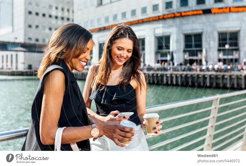 Zwei Frauen auf einer Brücke mit Kaffee zum Mitnehmen und Mobiltelefon lächeln Kollegin Kolleginnen Handy Handies Handys Mobiltelefone weiblich Getränk Getraenk