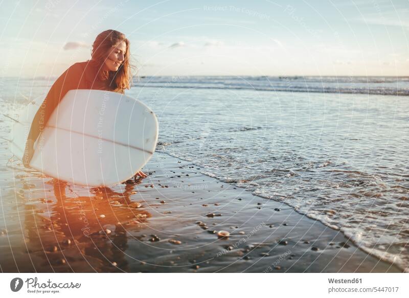 Junge Frau mit Surfbrett kauert am Meeresufer Surfbretter surfboard surfboards Surferin Wellenreiterinnen Surferinnen Surfen Surfing Wellenreiten Wassersport