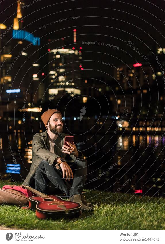 Lächelnder junger Mann mit Gitarre und Handy sitzt nachts am städtischen Flussufer Mobiltelefon Handies Handys Mobiltelefone Männer männlich lächeln Nacht