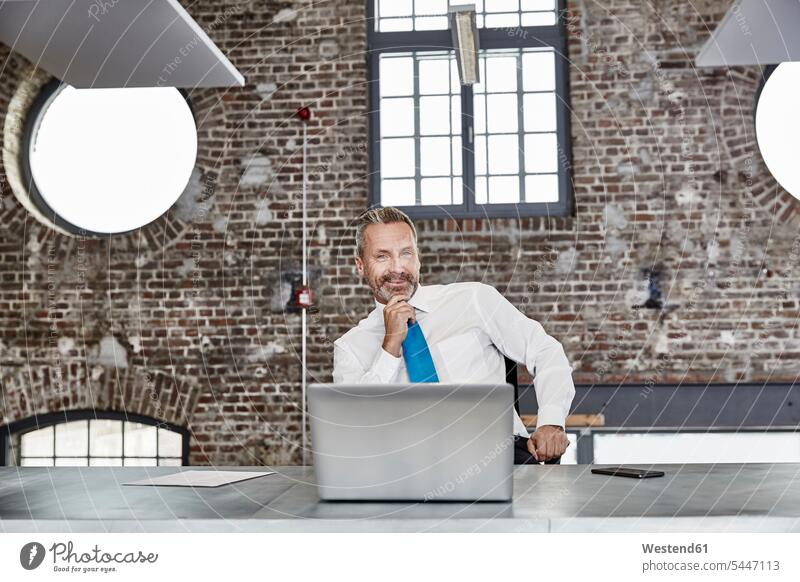 Porträt eines selbstbewussten Geschäftsmannes mit Laptop, der in einem Loft am Tisch sitzt sitzen sitzend Notebook Laptops Notebooks lächeln Businessmann