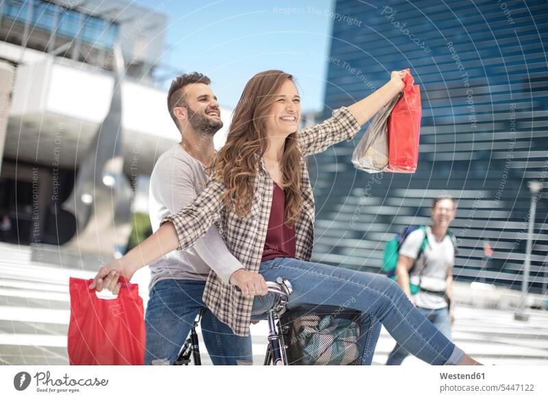 Glückliches junges Paar amüsiert sich in der Stadt beim Fahrradfahren mit Einkaufstaschen Spaß Spass Späße spassig Spässe spaßig Bikes Fahrräder Räder Rad