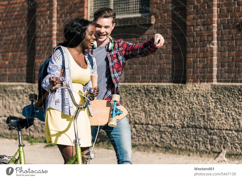 Junges Paar mit Fahrrad und Skateboard macht Smartphone-Selfies fröhlich Fröhlichkeit Frohsinn Heiterkeit multikulturell iPhone Smartphones Handy Mobiltelefon