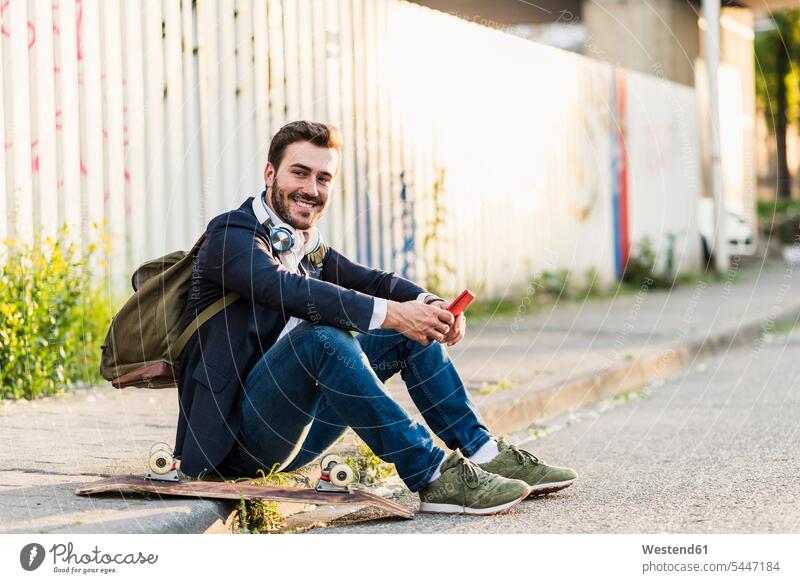 Lächelnder junger Mann sitzt auf dem Bürgersteig und hält ein Handy lächeln sitzen sitzend Männer männlich Mobiltelefon Handies Handys Mobiltelefone Erwachsener