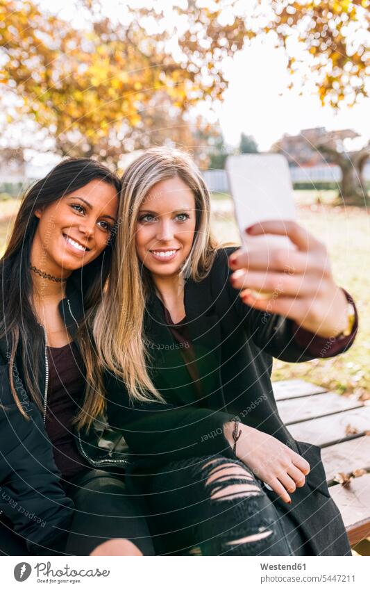 Zwei lächelnde junge Frauen auf einer Parkbank machen ein Selfie Selfies weiblich Parkanlagen Parks Bank Sitzbänke Bänke Sitzbank Freundinnen Handy Mobiltelefon