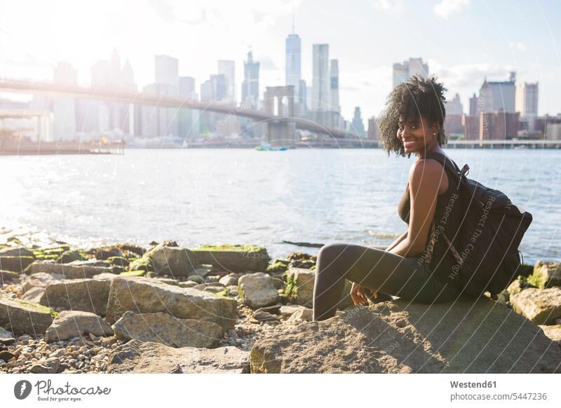 USA, New York City, Brooklyn, Porträt einer lächelnden Frau, die an der Uferpromenade sitzt weiblich Frauen Skyline Skylines Brücke Bruecken Brücken sitzen