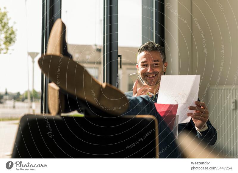 Lächelnder Geschäftsmann sitzt mit erhobenen Füßen in seinem Büro und benutzt Smartphone und Kopfhörer telefonieren anrufen Anruf telephonieren Businessmann