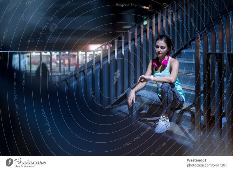 Junge Frau mit rosa Kopfhörern sitzt auf der Treppe und schaut auf ihre Smartwatch Joggerin Joggerinnen Uhrzeit checken trainieren junge Frau junge Frauen