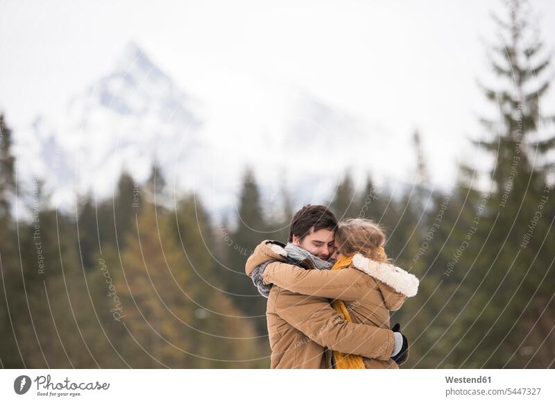 Glückliches junges Paar umarmt sich in Winterlandschaft Pärchen Paare Partnerschaft winterlich Winterzeit umarmen Umarmung Umarmungen Arm umlegen Mensch
