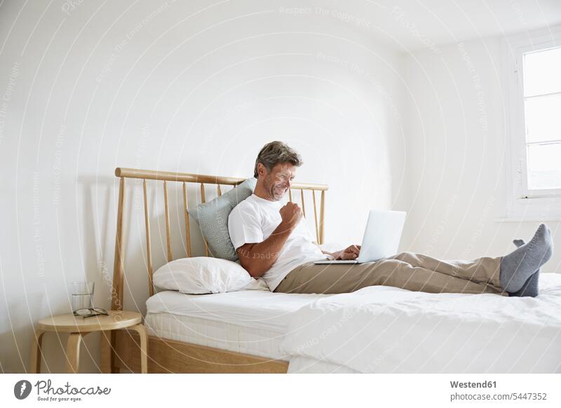 Reifer Mann genießt Erfolg mit Laptop im Bett Betten lächeln Notebook Laptops Notebooks Schlafzimmer Zuhause zu Hause daheim Männer männlich jubeln Jubel sitzen