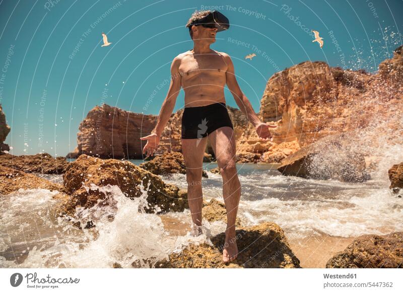 Erwachsener Mann mit VR-Brille zwischen Felsen am Strand Brillen Virtuelle Realität Virtuelle Realitaet Männer männlich Beach Straende Strände Beaches Gestein