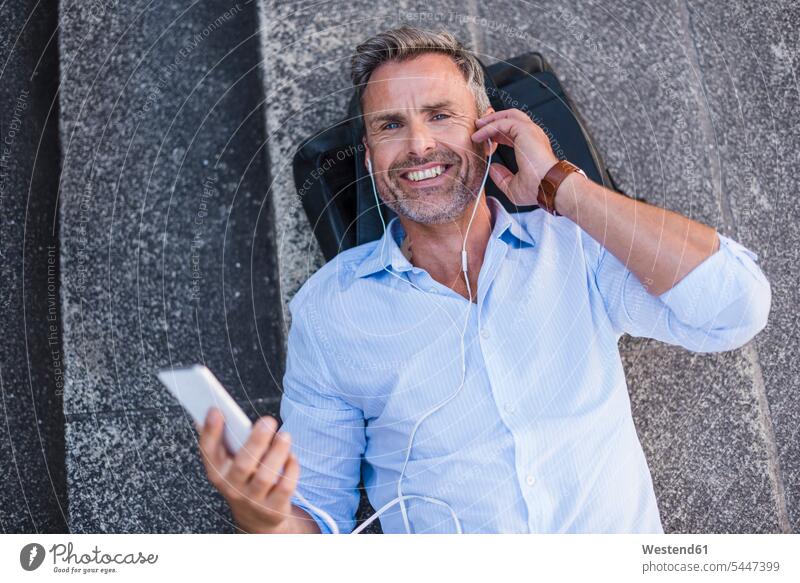 Lächelnder Mann mit Handy und Ohrstöpseln auf der Treppe liegend liegt lächeln Ohrhörer entspannt entspanntheit relaxt Mobiltelefon Handies Handys Mobiltelefone