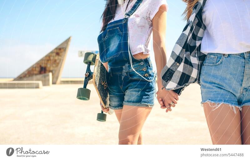 Zwei junge Frauen mit Longboards gehen Hand in Hand auf der Strandpromenade, Teilansicht Sexy Hot Pants Textfreiraum Jeans-Shorts Spanien Skateboard Rollbretter