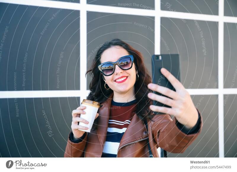 Porträt einer lächelnden jungen Frau mit Kaffee zum Mitnehmen, die ein Selfie mit ihrem Smartphone macht Selfies weiblich Frauen Erwachsener erwachsen Mensch