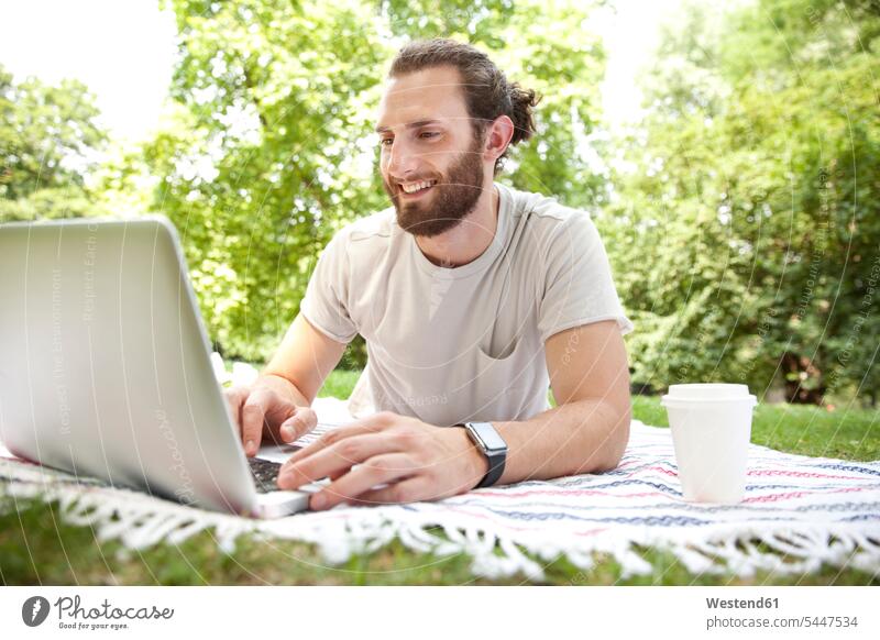 Lächelnder Mann mit Kaffee zum Liegen auf einer Decke in einem Park mit Laptop Notebook Laptops Notebooks Männer männlich Computer Rechner Erwachsener erwachsen