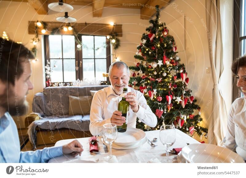 Lächelnder älterer Mann mit Familie hält eine Flasche Wein am Weihnachtstisch Weihnachten Christmas X-Mas X mas Weine Familien lächeln feiern Feste Festtag