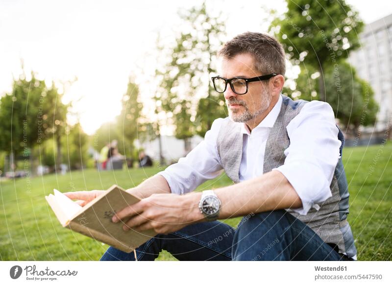 Selbstbewusster reifer Geschäftsmann im Stadtpark sitzt auf Gras und liest Buch Wiese Wiesen Bücher lesen Lektüre entspannt entspanntheit relaxt Mann Männer
