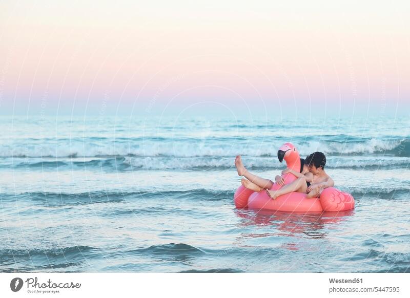 Verliebtes Paar schwebt mit aufblasbarem rosa Flamingo bei Sonnenuntergang auf dem Meer Meere Pärchen Paare Partnerschaft küssen Küsse Kuss Gewässer Wasser