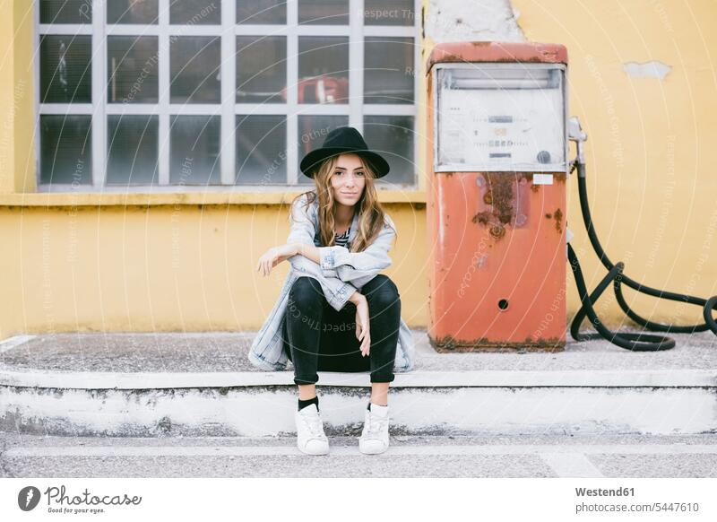 Porträt einer modischen jungen Frau mit Hut, die neben einer alten Zapfsäule sitzt Hüte Tanksäule Kraftstoffpumpe Zapfsaeulen Tanksaeulen Kraftstoffpumpen