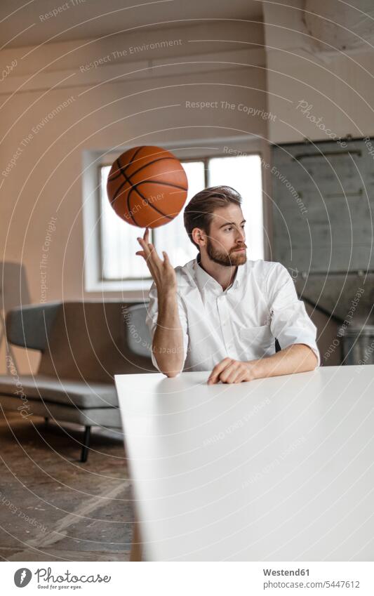 Porträt eines nachdenklichen jungen Geschäftsmannes, der in einem Loft am Tisch sitzt und Basketball auf seinem Finger balanciert Businessmann Businessmänner