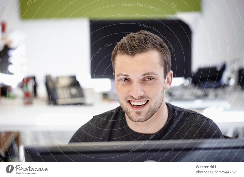 Junger Mann arbeitet im Büro und schaut in die Kamera sitzen sitzend sitzt Jungunternehmer arbeiten Arbeit Portrait Porträts Portraits lächeln Computer Rechner