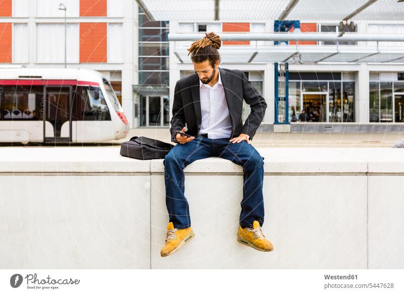 Junger Geschäftsmann mit Dreadlocks benutzt Smartphone beim Warten am Bahnhof Businessmann Businessmänner Geschäftsmänner iPhone Smartphones Geschäftsleute