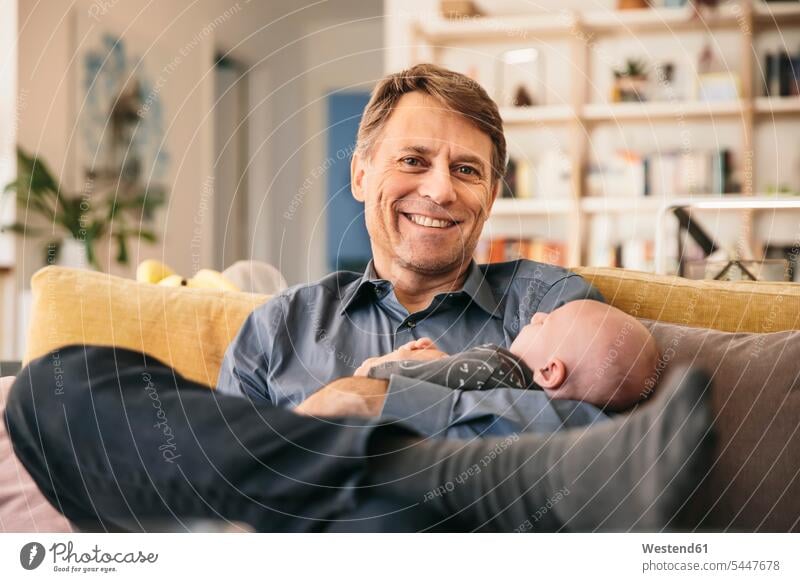 Lächelnder Vater und sein Neugeborenes sitzen zu Hause auf der Couch Sofa Couches Liege Sofas Papas Väter Vati Vatis Papis Kind Babies Babys Kinder lächeln