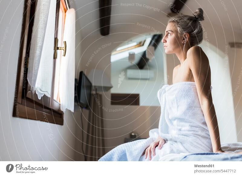 In ein Handtuch gewickelte junge Frau sitzt auf dem Bett und schaut aus dem Fenster weiblich Frauen schauen sehend sitzen sitzend Handtuecher Handtücher Betten