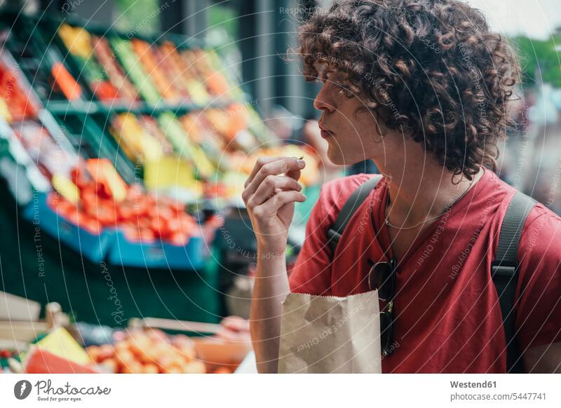 Junger Mann isst Kirschen vor einem Straßenobststand Männer männlich Obststand Obststaende Obststände essen essend Erwachsener erwachsen Mensch Menschen Leute